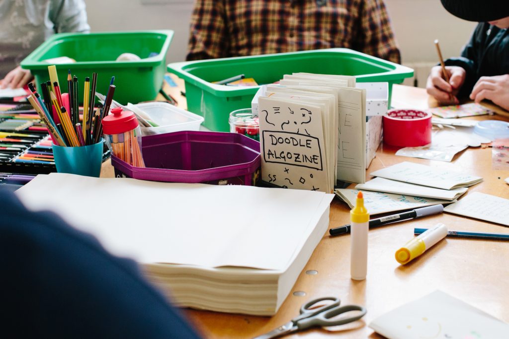 A table with paper, coloured pencils, and a folded piece of paper which reads 'doodle algozine'