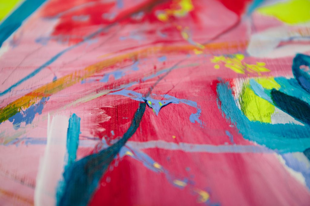 A close up of a painting with expressive layered brushstrokes. This closeup view is abstract and made up of bright blues, yellows, reds and pinks.