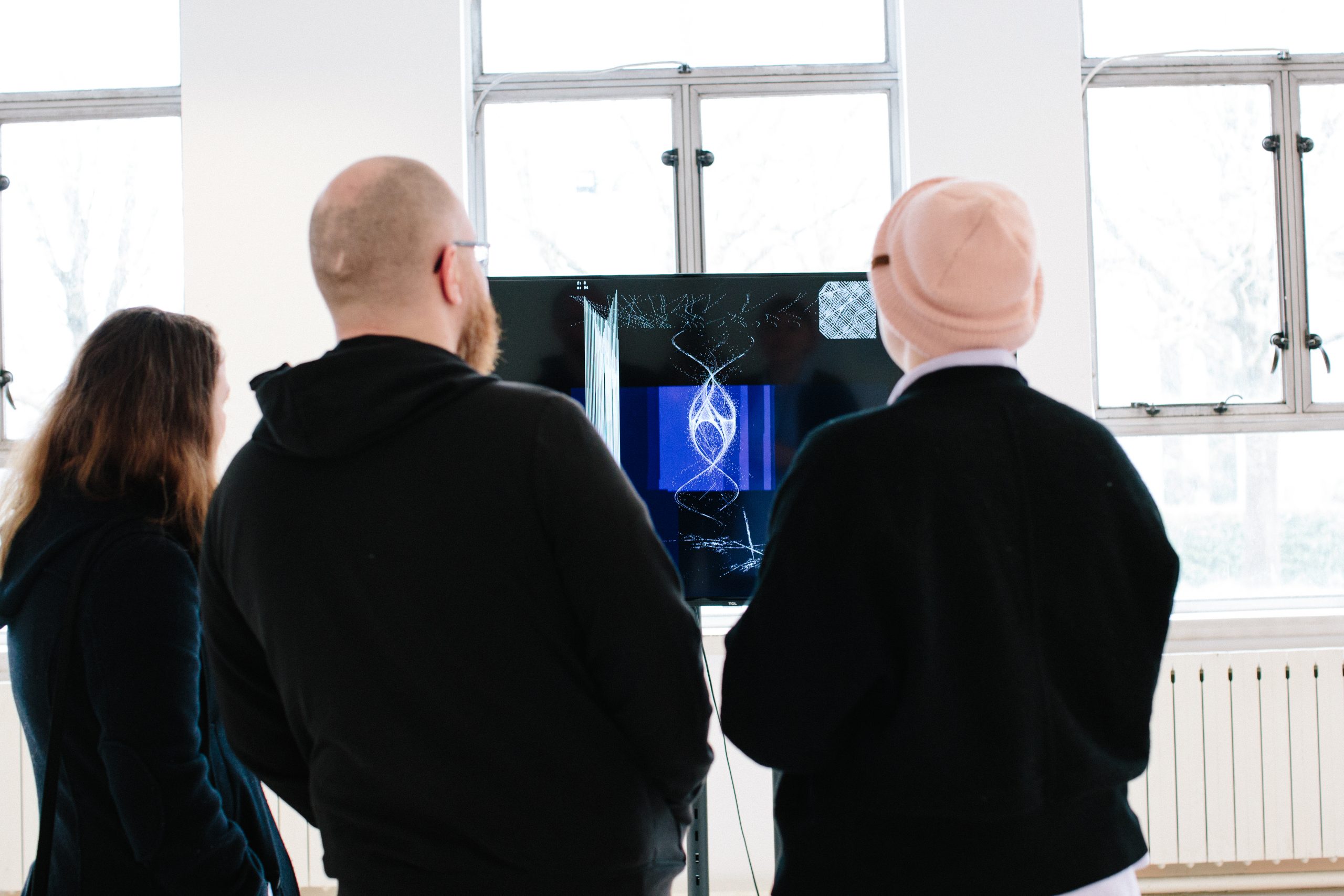 Three people from behind, observing an intricate digital art piece on a monitor in a bright, naturally lit room.
