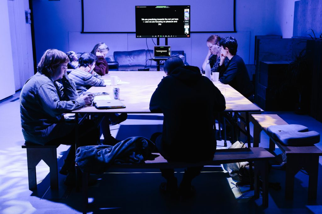 A group of people sat around a table in dim blue lighting, with a presentation on a screen in the background.