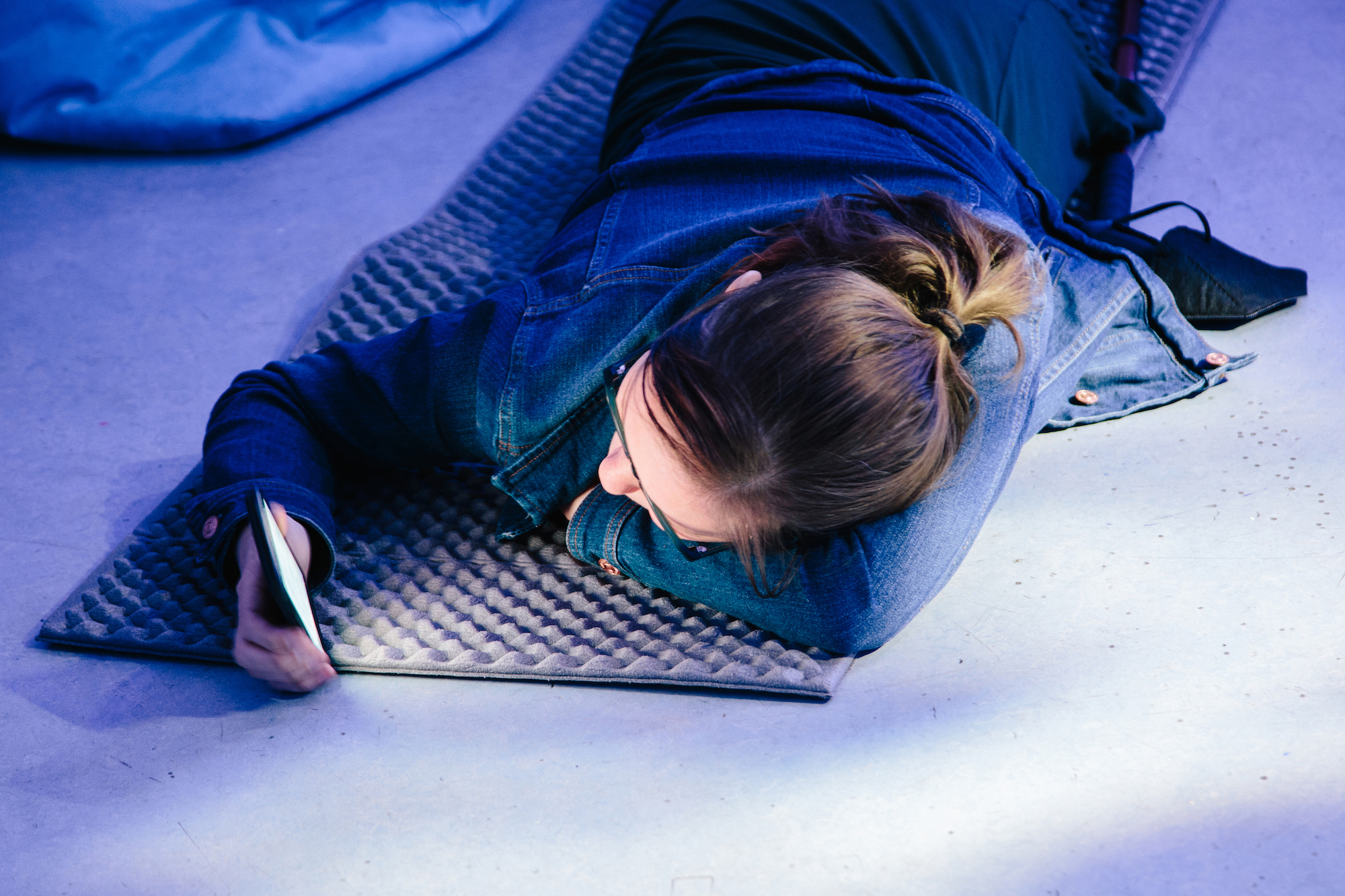 A person lies prone on a grey mat on the floor, resting their head on their arm, while looking at a glowing phone screen.