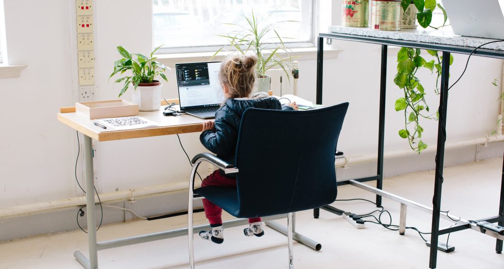 A child sits at a desk using a laptop in a room with large windows and potted plants.