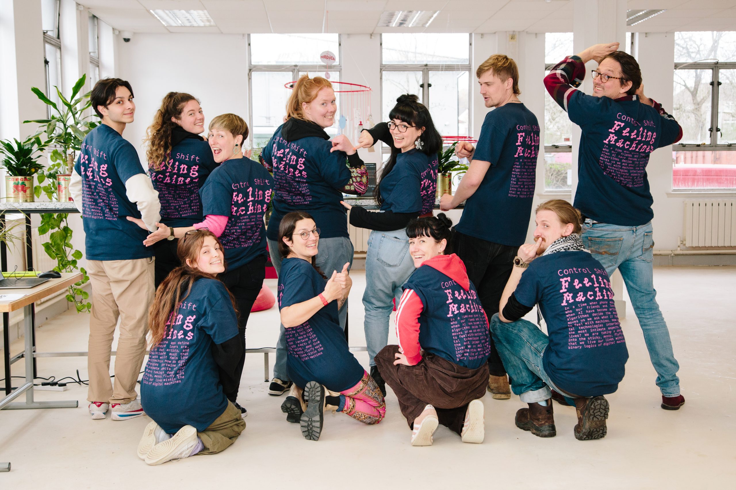 A group of people wearing matching dark blue t-shirts with pink text, playfully posing together. The shirts read 'Control Shift Feeling Machines'
