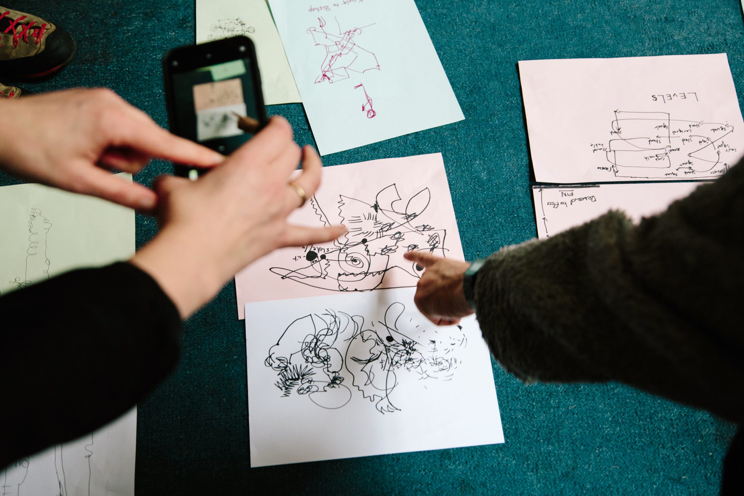 Close-up of hands pointing at and photographing abstract line drawings and diagrams on paper spread out on the floor.