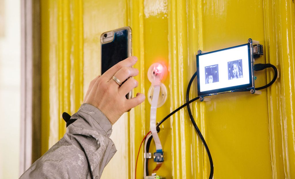 A person's hand holding a smartphone, capturing an electronic device with a small screen displaying images, mounted on a yellow wall.