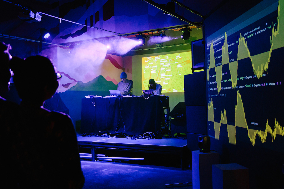 Two musicians on a stage looking at laptops, digital sound wave visuals are projected next to them.