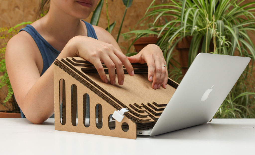 A cardboard structure which has been placed over an open laptop. A person rests their hand on the structure, which tilts at a 45 degree angle down towards the laptop.