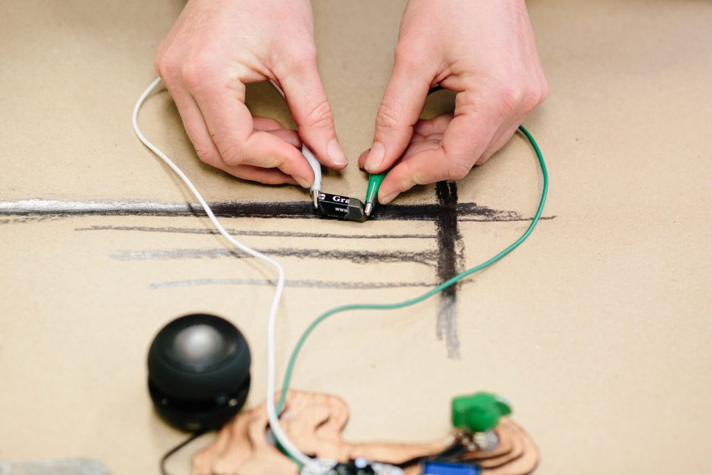 Close-up of hands connecting wires to an electronic device on a large sheet of paper with black lines drawn on it.