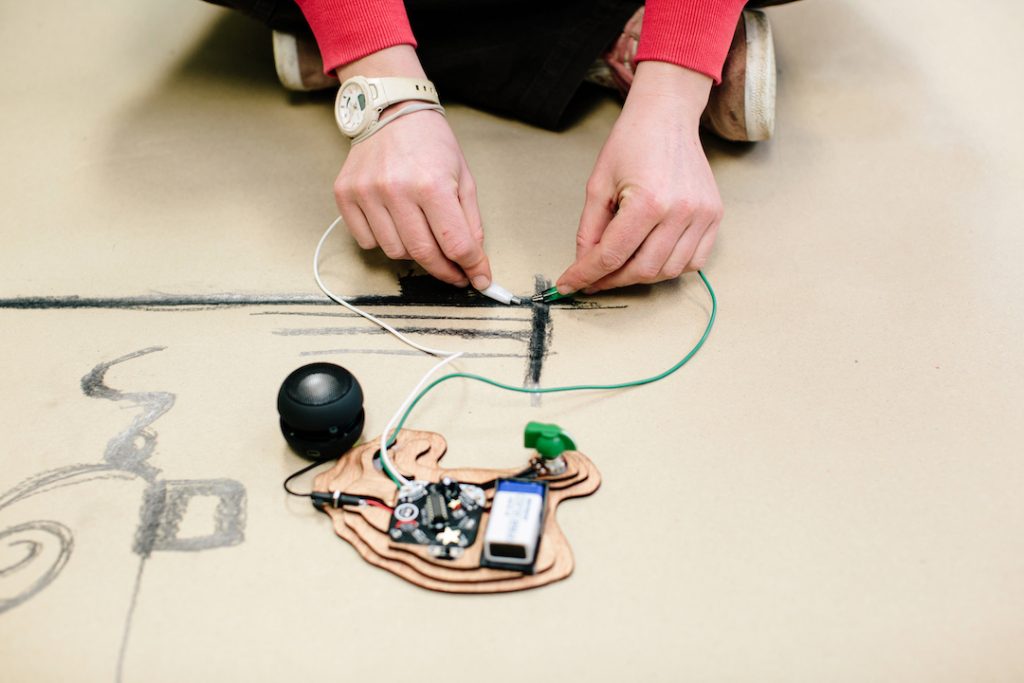 Close-up of hands connecting wires to an electronic device on a large sheet of paper with black lines drawn on it.