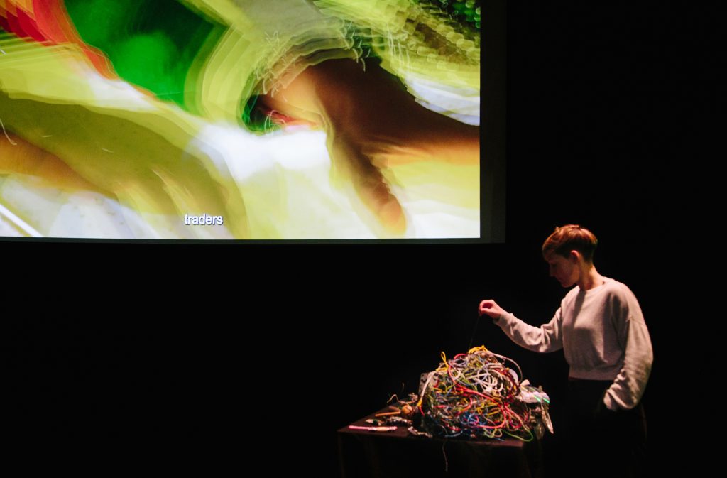 A person interacting with a pile of tangled wires on a table in front of them. Behind them is a colourful, abstract projection