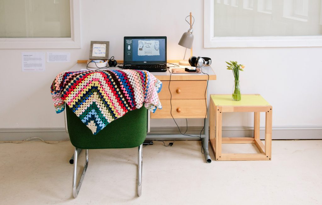 A green chair with a cosy blanket in front of a desk with a laptop, lamp, and vase of flowers.