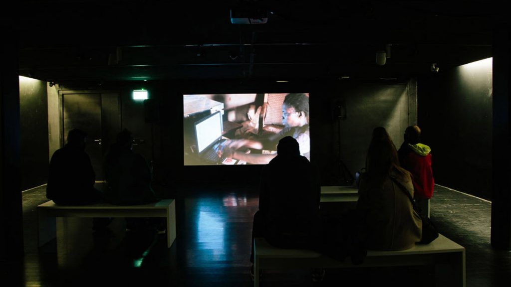 People sat on benches in a dark space, facing a projected film of a person typing on a computer.