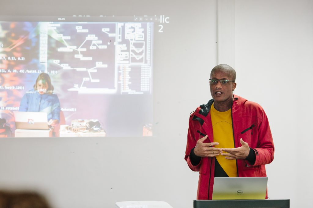 A speaker wearing a red jacket presenting in front of a screen showing digital art software.