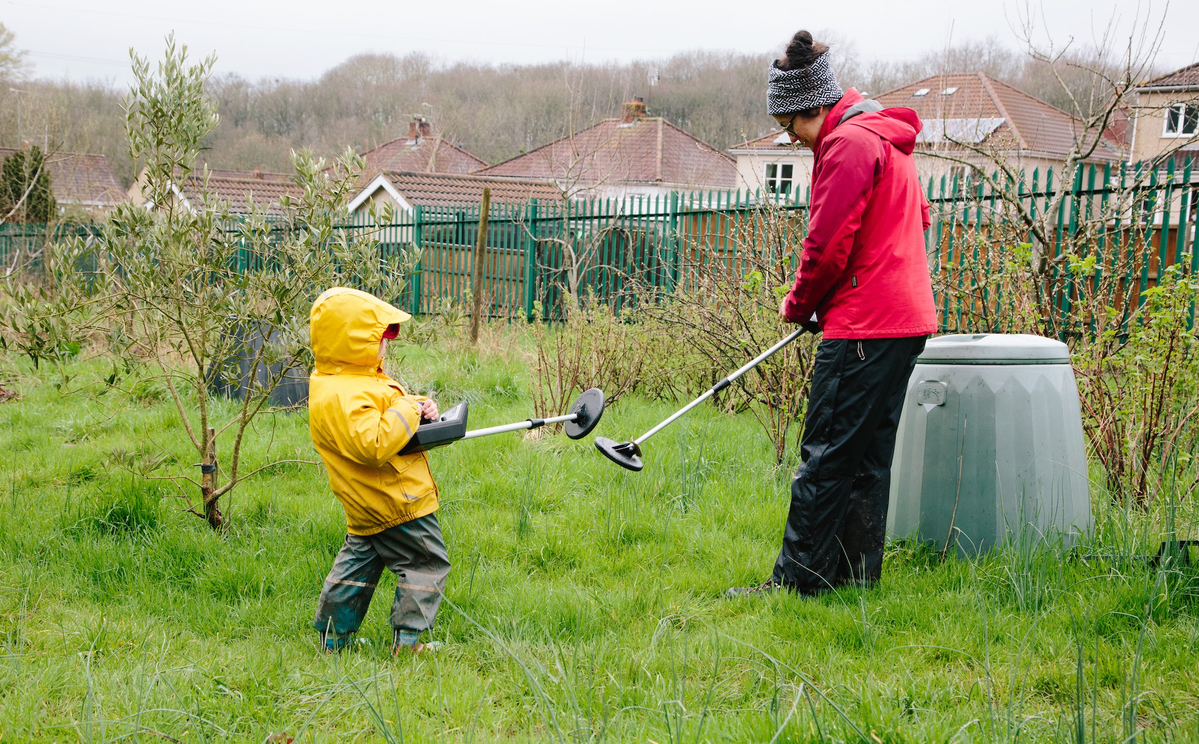 A child in a yellow raincoat and an adult in a red jacket using metal detectors on a grassy field with houses in the background.