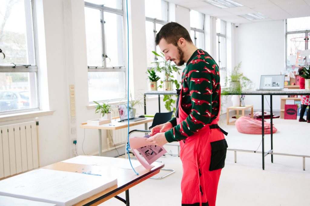 A person wearing red dungarees is reading a pink booklet at a workstation. The room is filled with natural light and potted plants.