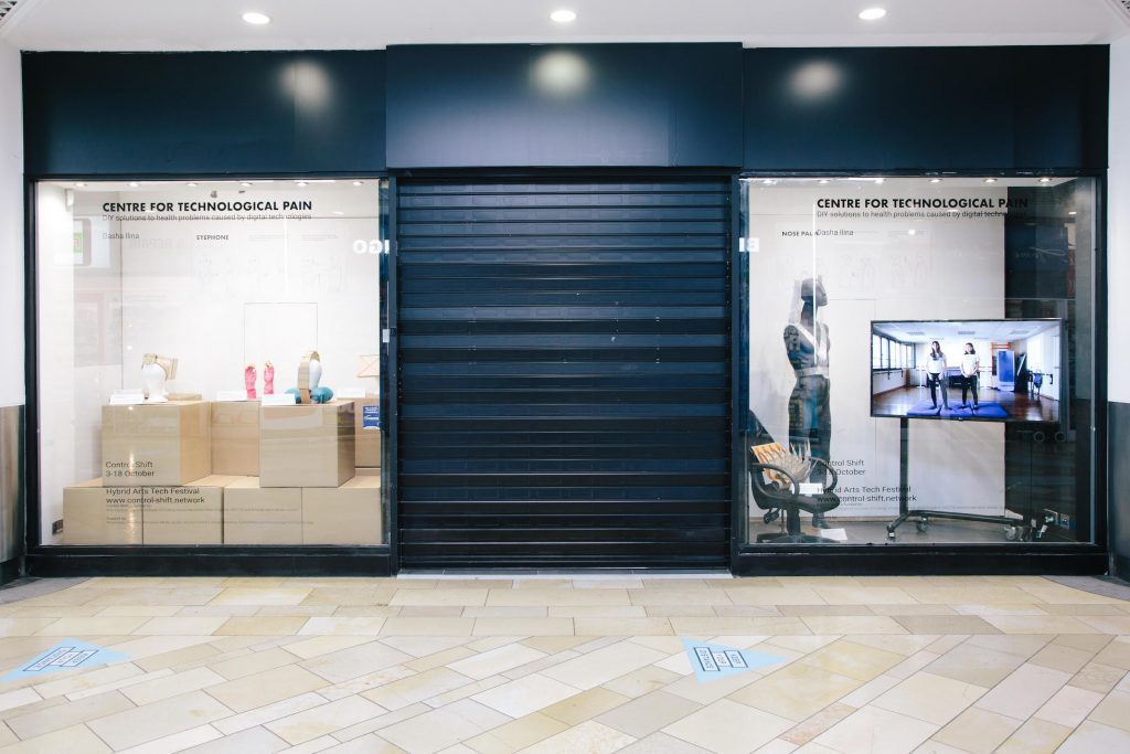 A window display inside a shopping mall. There is a back metal shutter covering the middle, with exhibitions either side showing mannequins wearing prototypes, a TV screen, and the words 'centre for technological pain' on the glass.