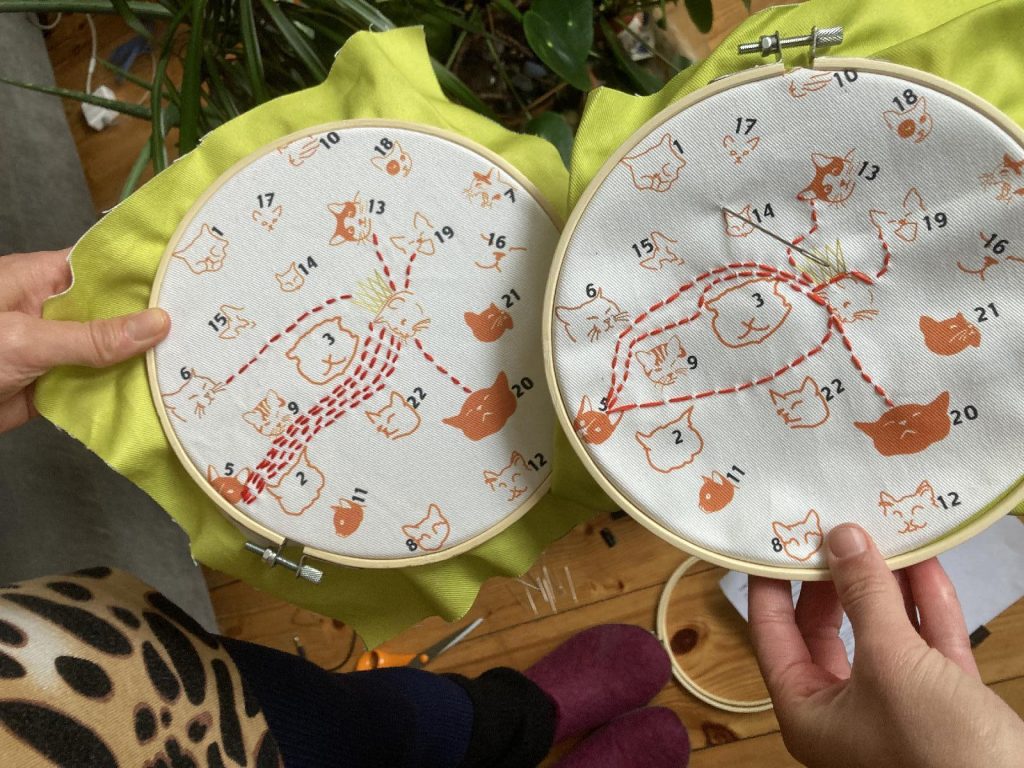 Two people holding their embroidery hoops together, both with cats and dotty lines and numbers.