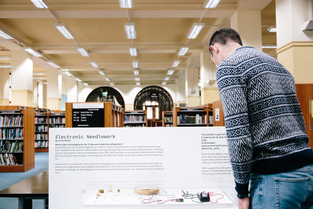 Inside Bristol Library, a person is viewing a glass cabinet filled with batteries, wires, and embroidery materials in front of a plaque which reads 'electronic needlework by Irene Posch.