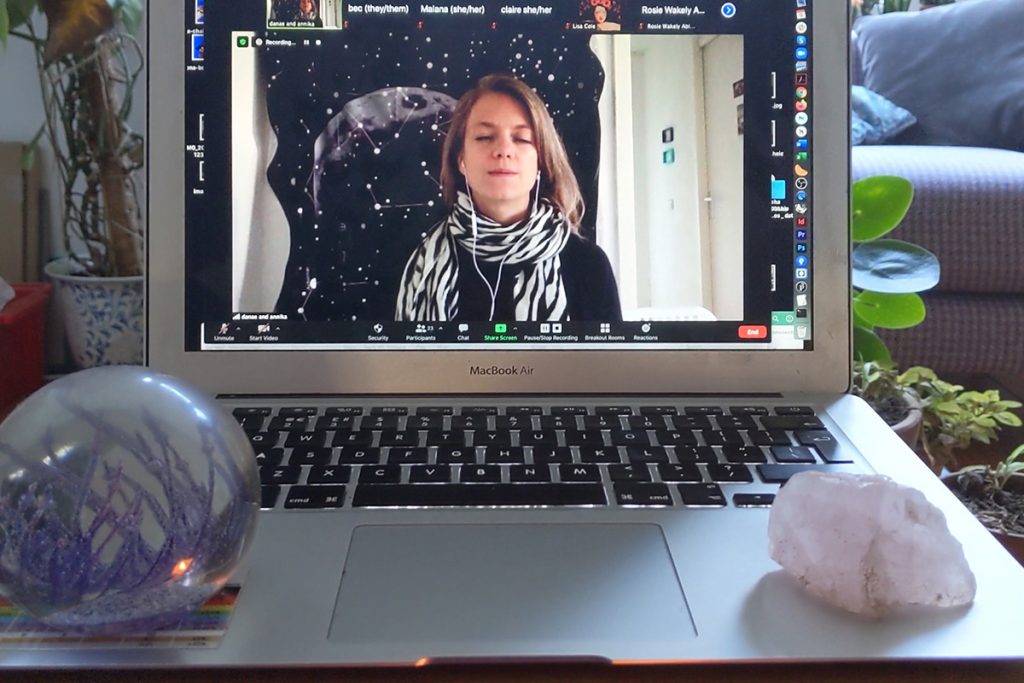 A laptop surrounded by plants and crystals. The screen shows a video caller with their eyes shut meditavely.