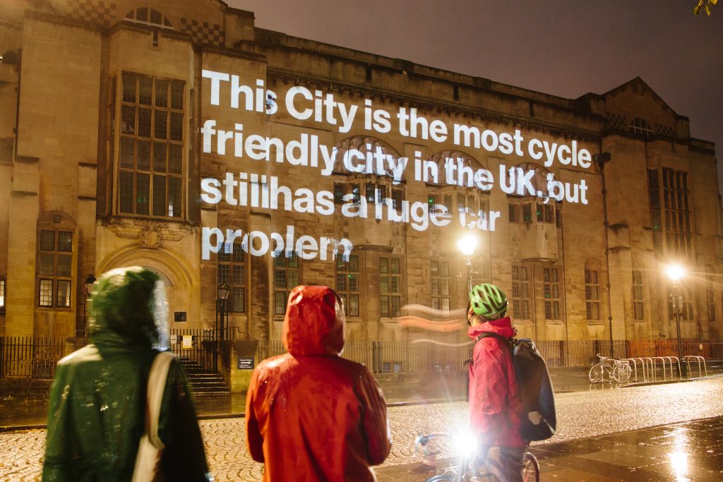 Projection of text onto a building which reads "This city is the most cycle friendly city in the UK, but still has a huge car problem"