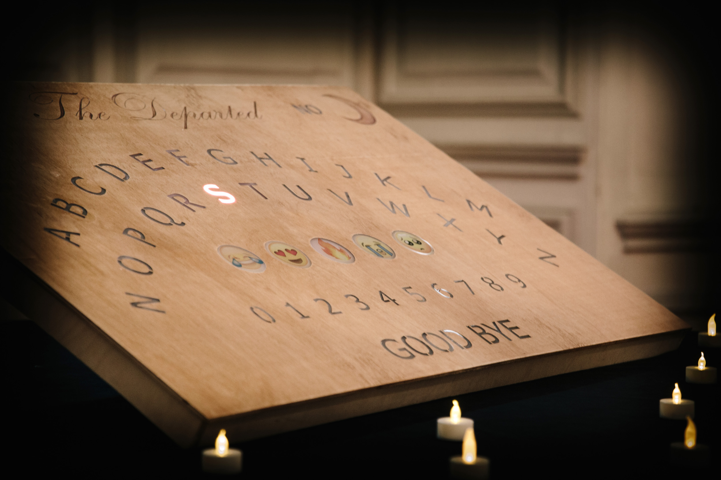 Close up of a wooden ouija board with the alphabet laid out in an arch, numbers 0 to 9, five emojis, and 'goodbye' written at the bottom. The letter 'S' is illuminated red.